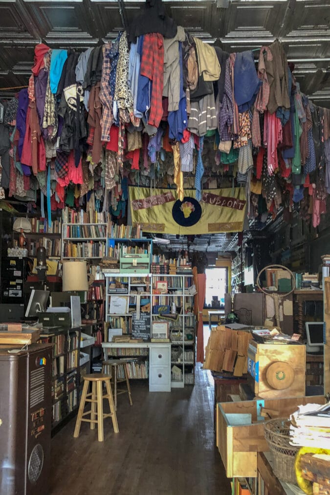 clothes hang from the ceiling of a shop packed with books, boxes and other items