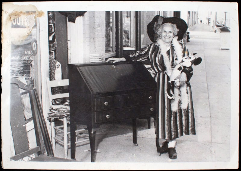 a black and white photo of a woman in a floppy hat, striped dress holding a mickey doll and smiling next to a piece of furniture on the sidewalk
