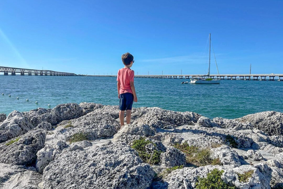 a boy in shorts and a red t-shirt stands on gray rocks and looks out at a sailboat on the blue water