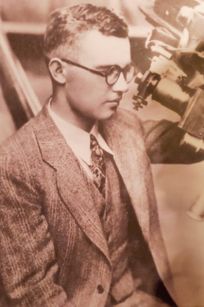 A black and white photo of a man wearing glasses and a suit looking through a telescope