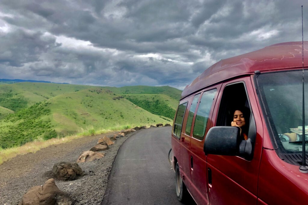 a maroon van on a road under cloudy skies with a woman in the passenger seat smiling