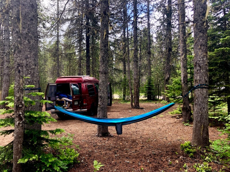 a red van is parked in the woods with a blue hammock strung up between two trees
