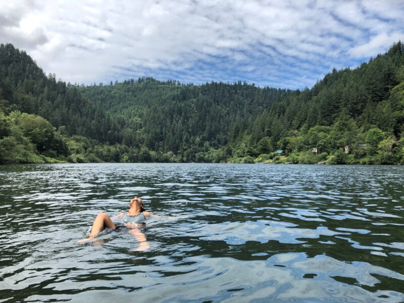 a person floats in a lake surrounded by blue skies and green pine trees