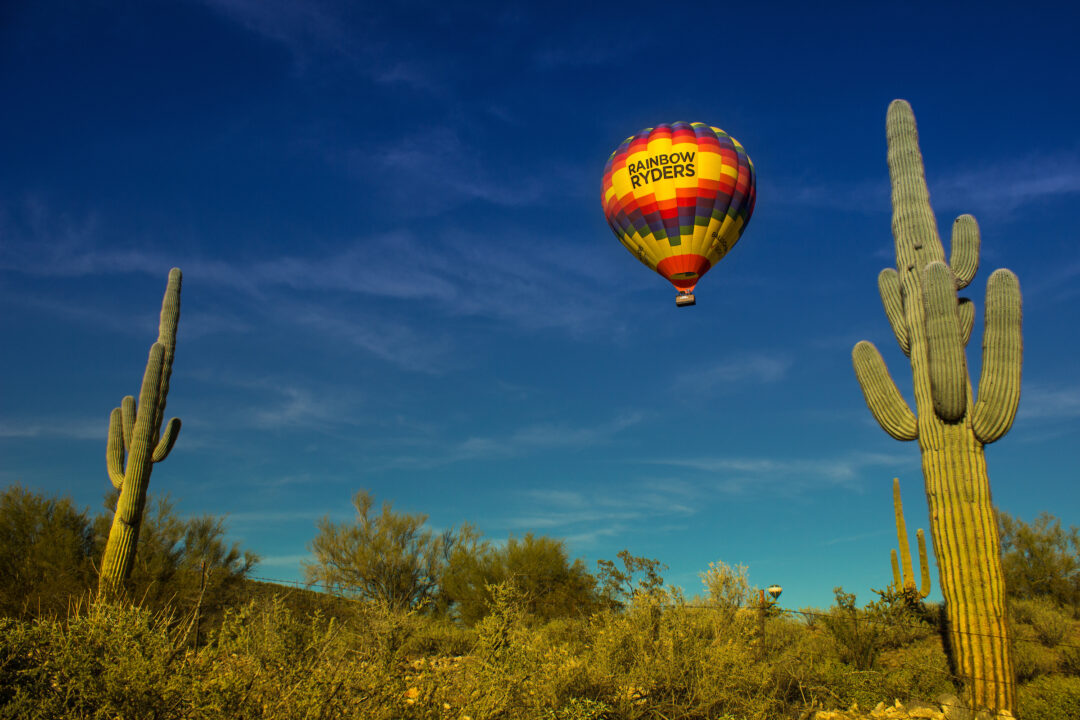 a red, yellow and blue hot air balloon flies over the desert and large cacti