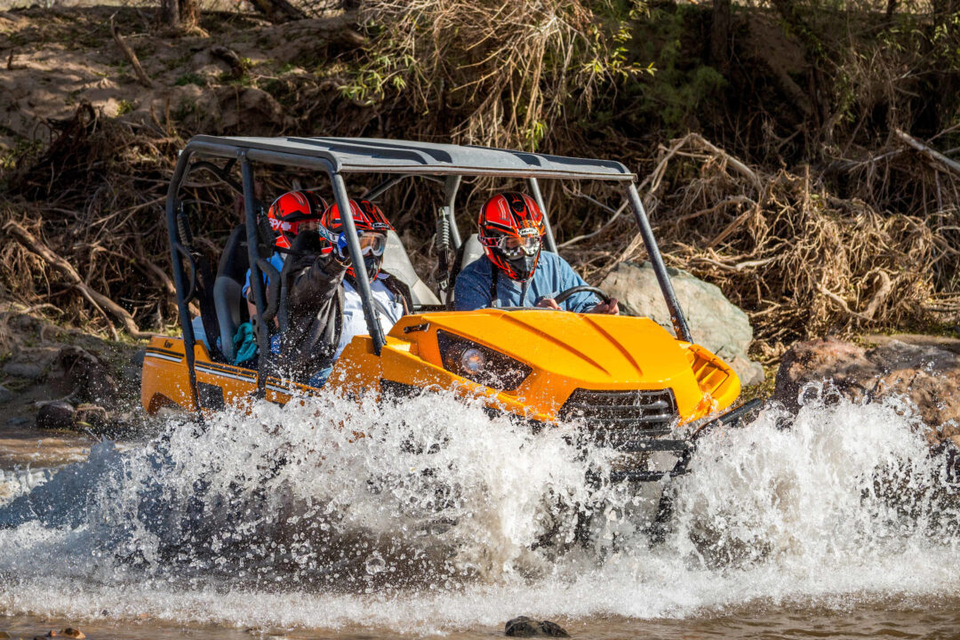 three people in red helmets drive an ATV through the water