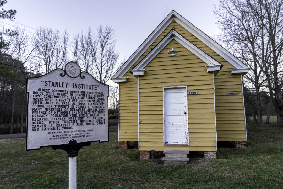 a white and yellow wooden one-room schoolhouse with a historic marker out front