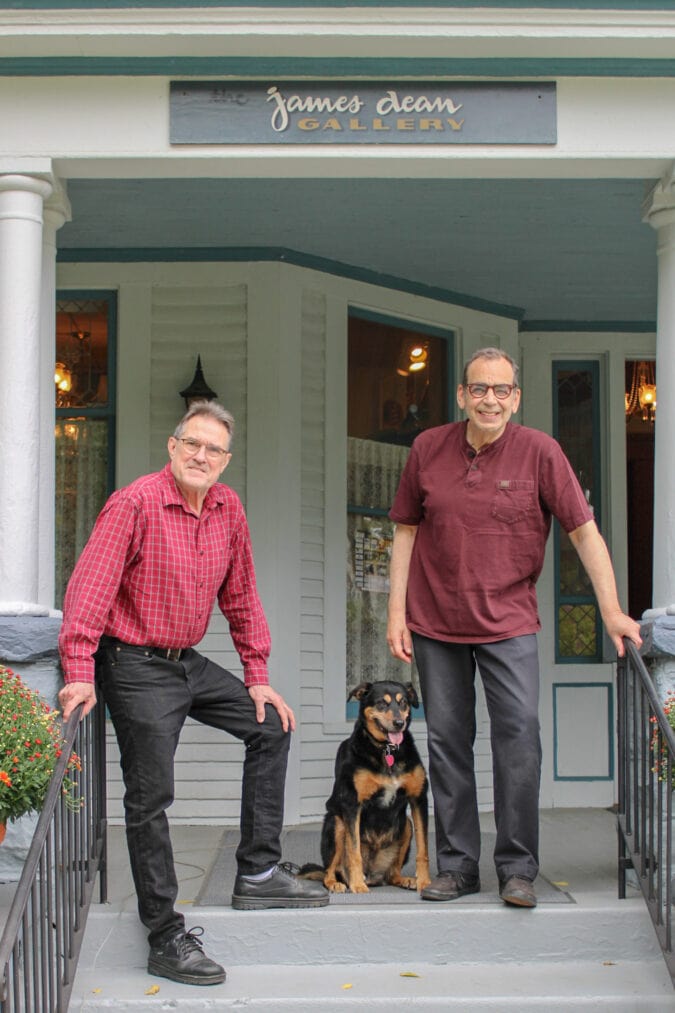 two men and a dog stand and smile on the porch of a victorian house under a sign that says "james dean gallery"