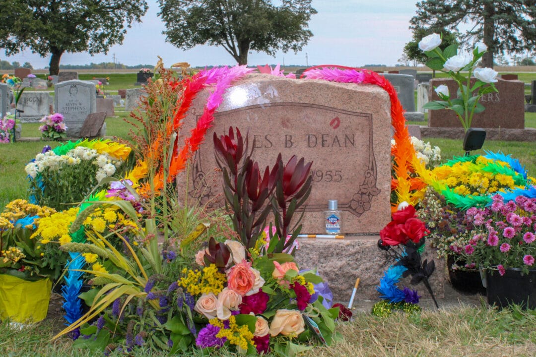 a graveyard with a pink marble headstone for james b dean covered in lipstick prints and draped in colorful flowers