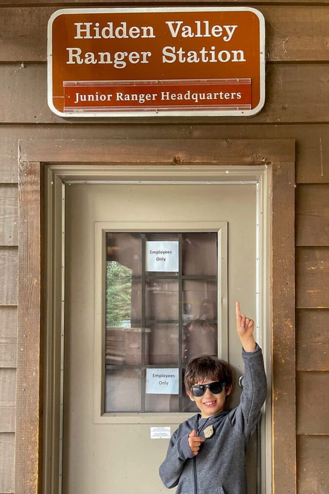 a boy in sunglasses smiles in front of a door and points to a sign that says "hidden valley ranger station, junior ranger headquarters."