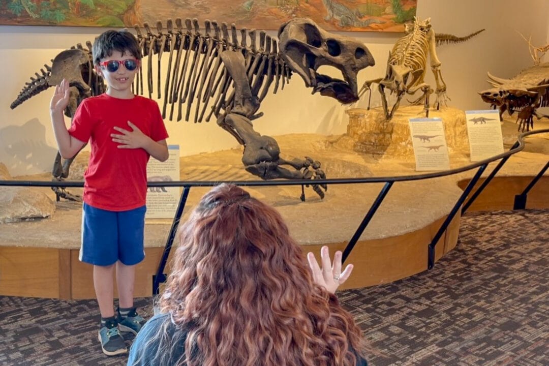 a boy raises one hand in front of a dinosaur skeleton and takes an oath from a park ranger