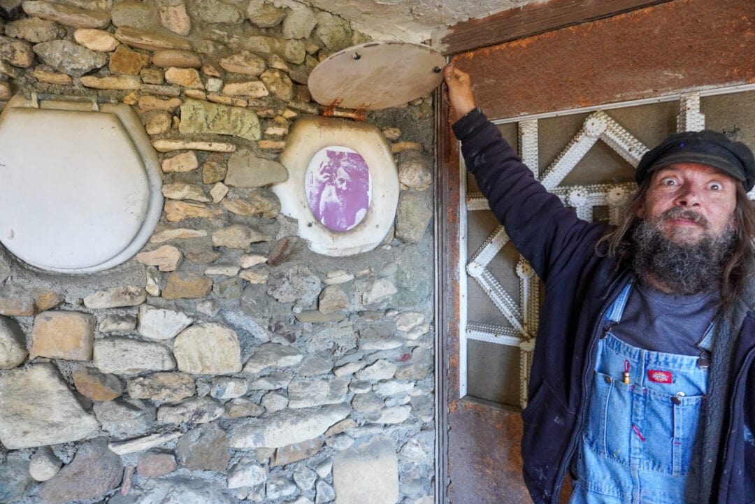 A man is holding up the lid of a toilet seat mounted to the wall, revealing a photo underneath