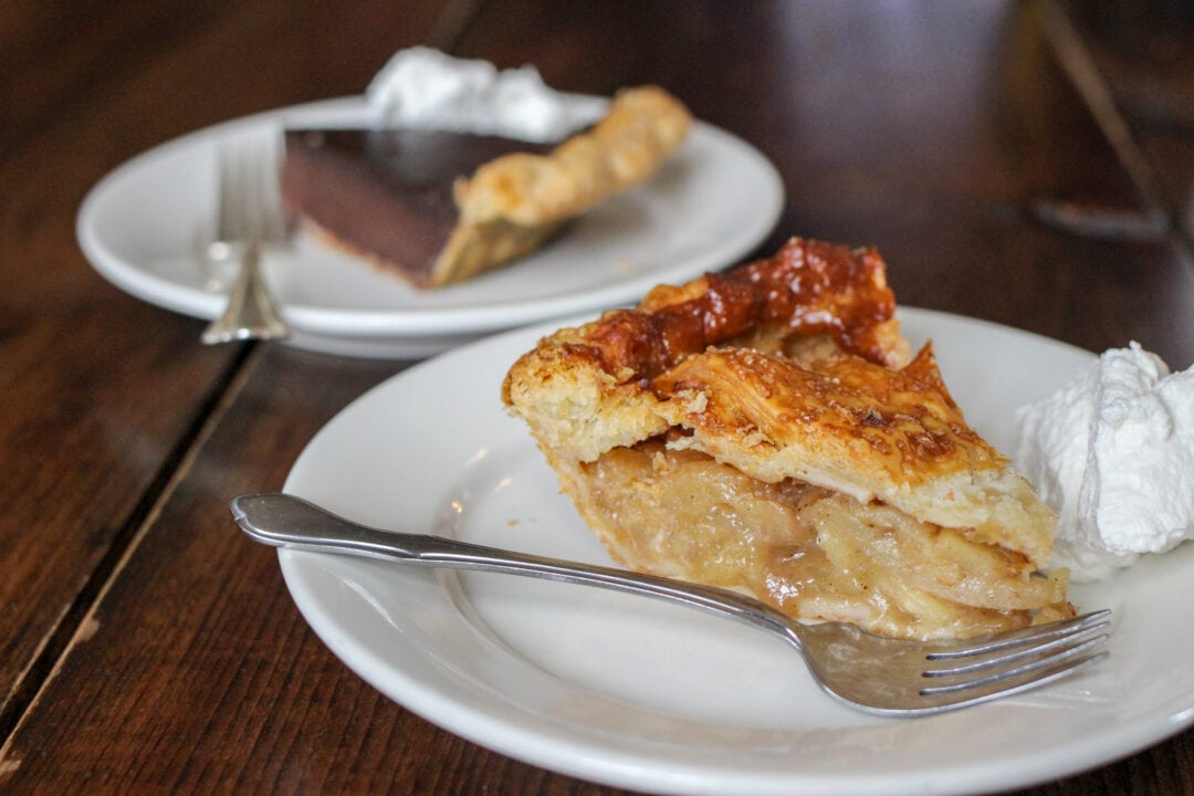 two slices of pie, one chocolate and one apple, sit on white plates with forks on a wooden table