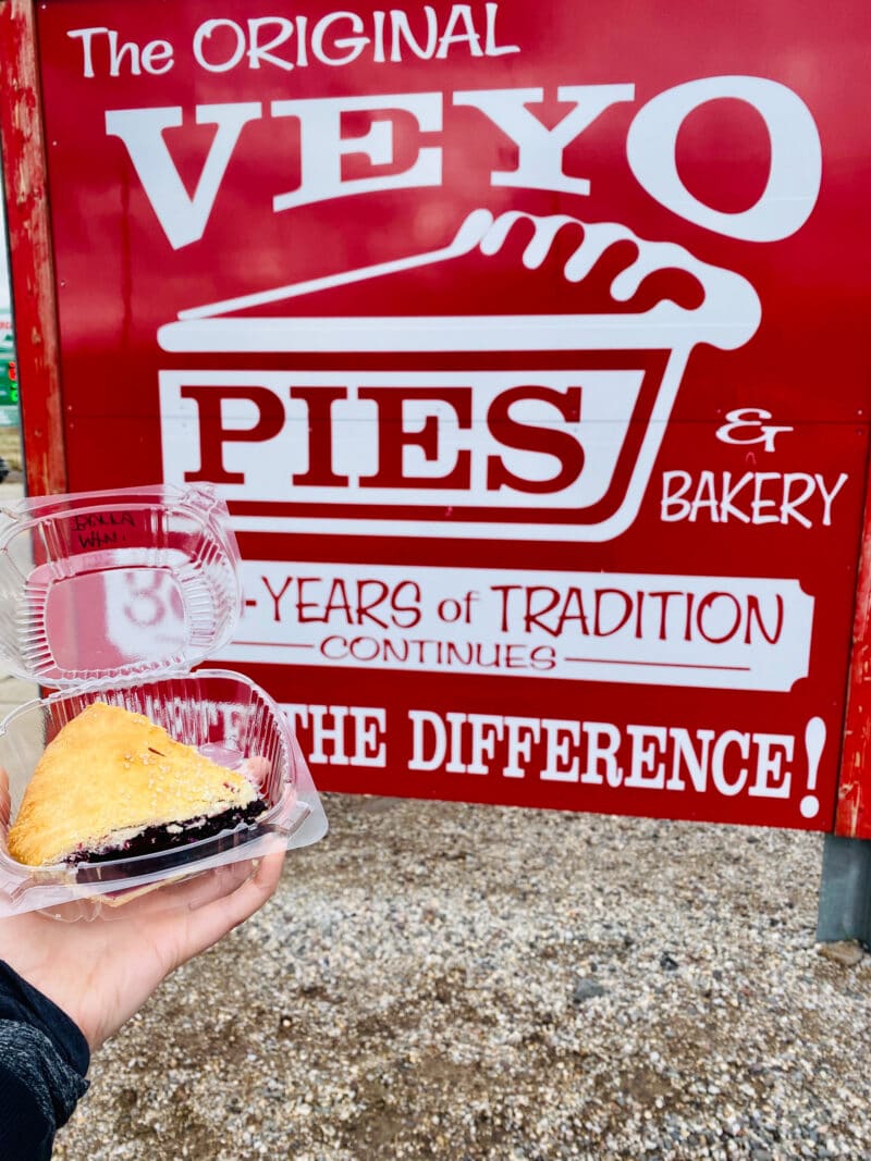 a hand holds a slice of pie in a plastic clamshell container in front of a red and white sign that says "the original veyo pies"