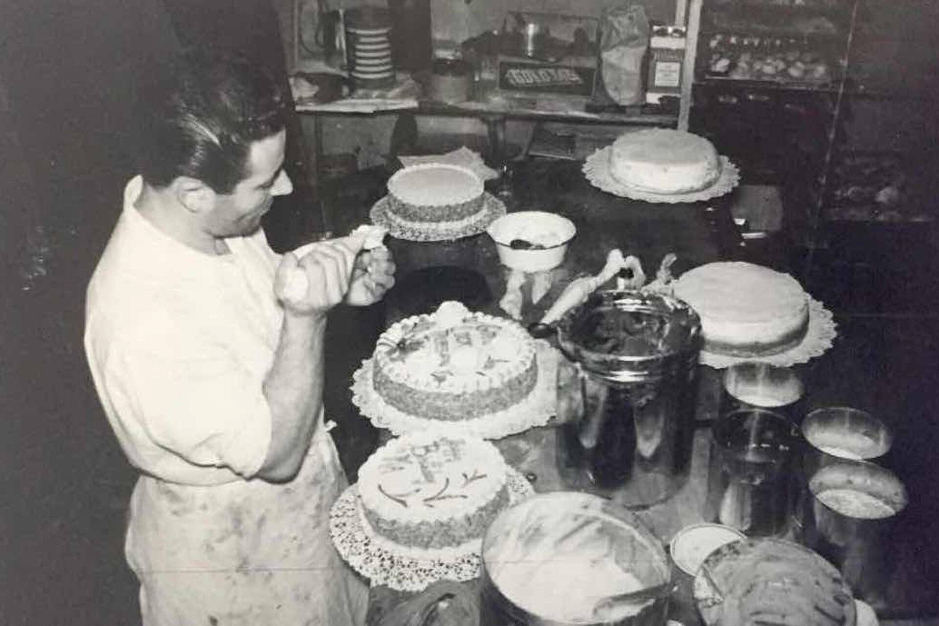 a black and white photo of a man in a white apron piping icing onto cakes