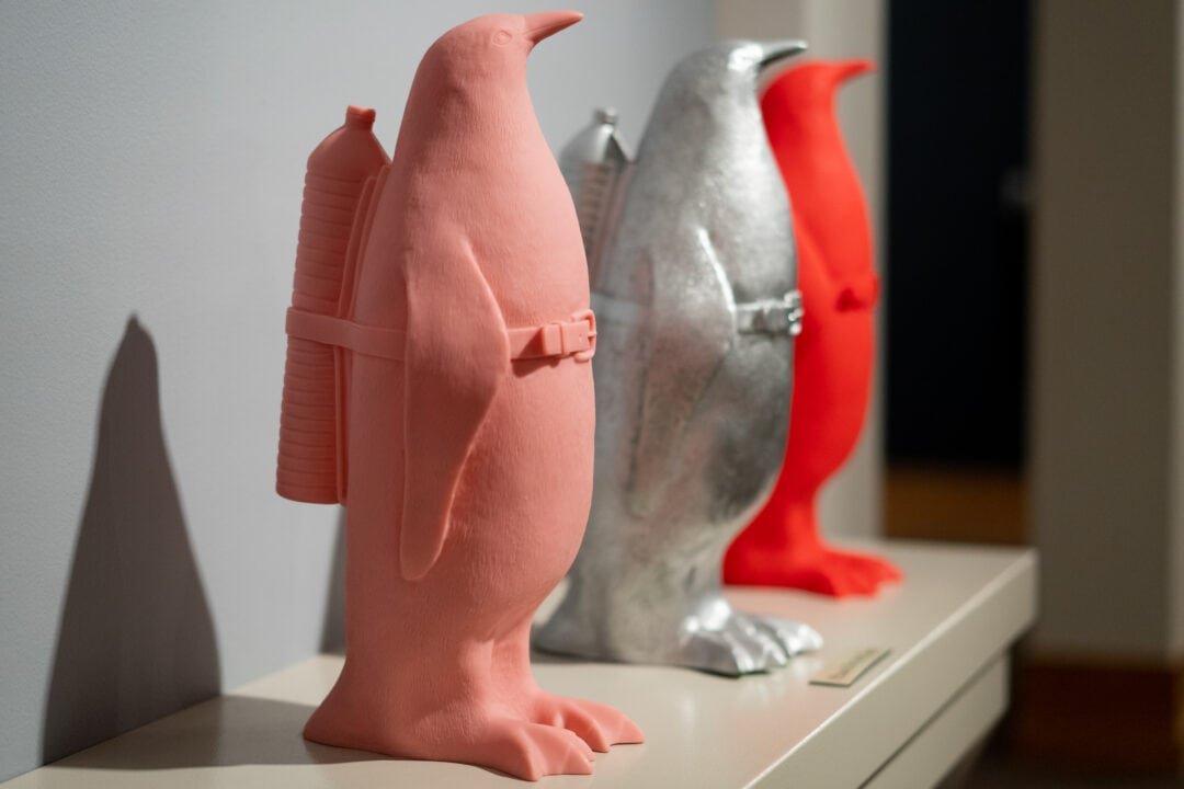 three plastic penguins, one pink, one silver, and one red, with water bottles strapped to their backs