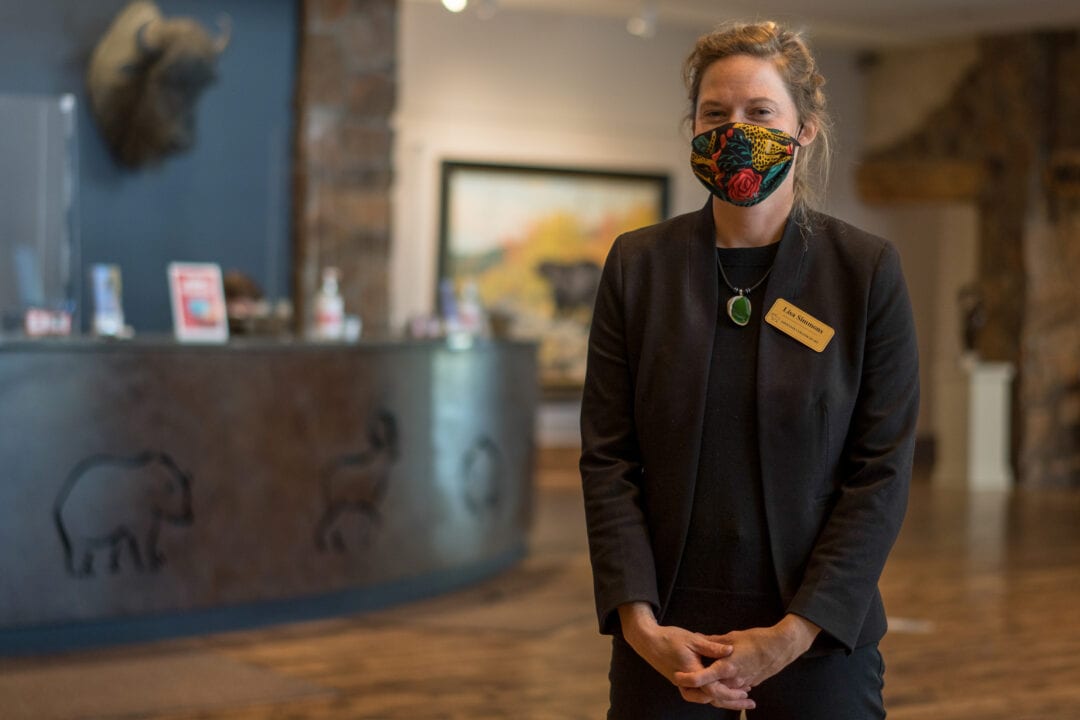 A woman wearing a face mask clasps her hand an poses for the camera in front of a museum front desk and buffalo head mount