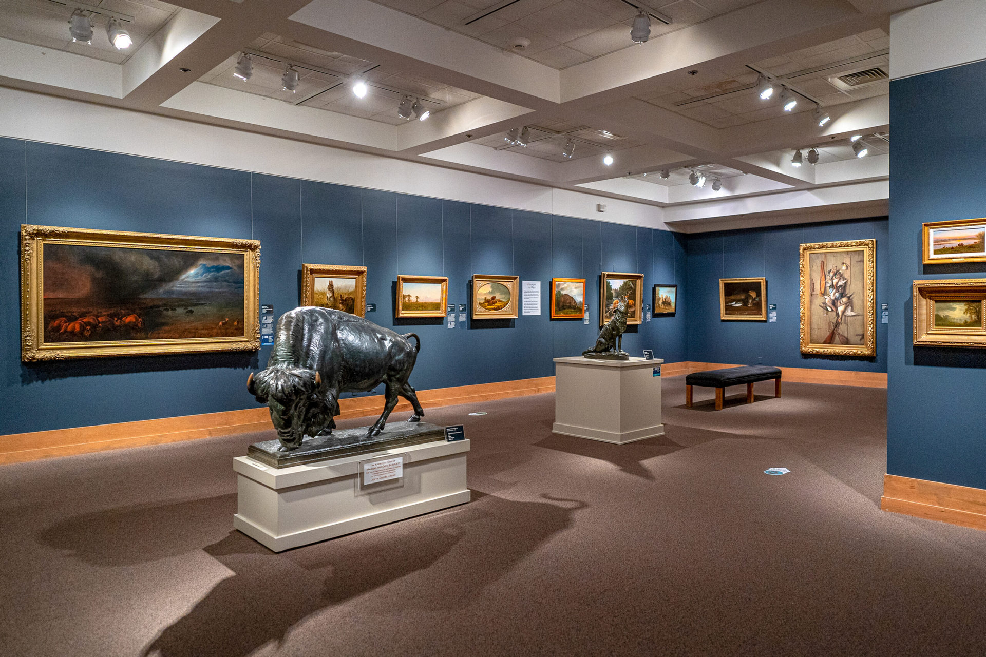 A gallery in an art museum featuring framed paintings hung on a blue wall and animal sculptures in the center of the well-lit room