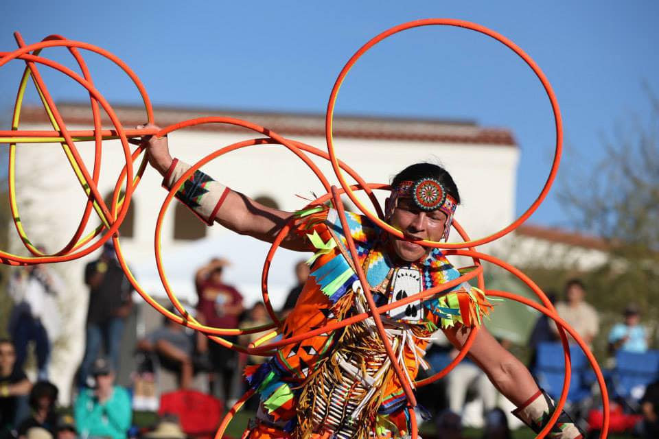 a native american hoop dancer dances with several orange hoops in front of a crowd outside