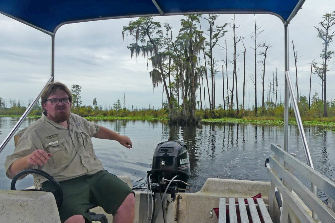 a man in a khaki shirt and shorts drives a pontoon boat on a body of water