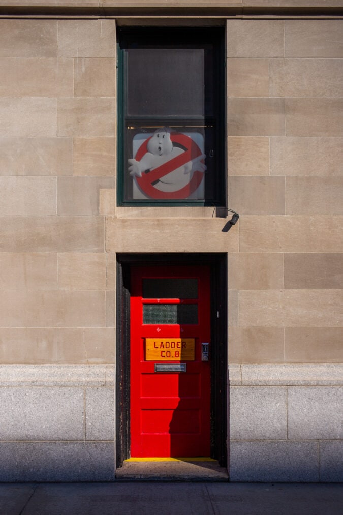a ghostbusters sign hangs in a window above a bright red door in a stone building