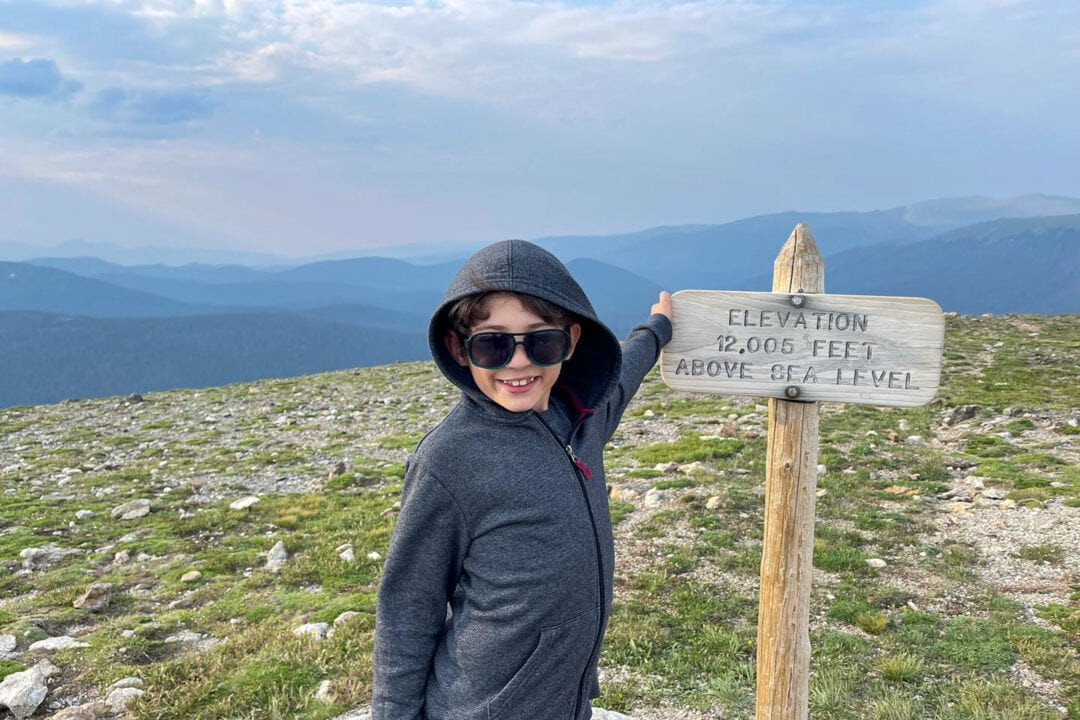 a kid in sunglasses and a hooded sweatshirt stands by a sign that reads "elevation 12,005 feet above sea level"