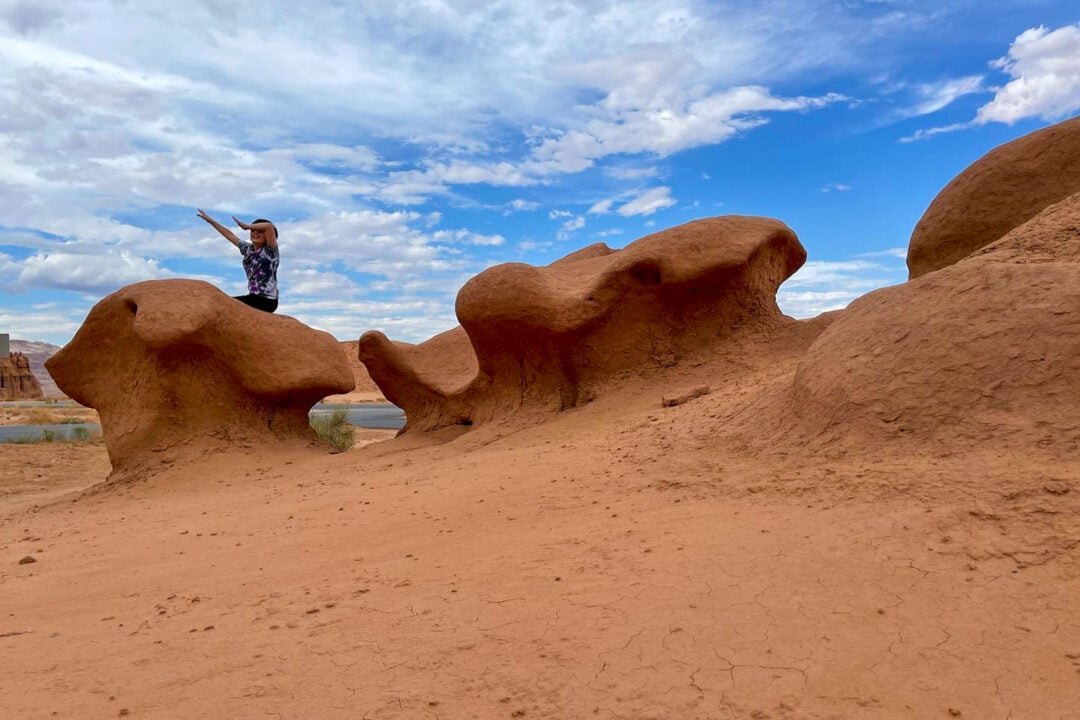 a kid sits on a red rock formation in front of a blue sky with clouds