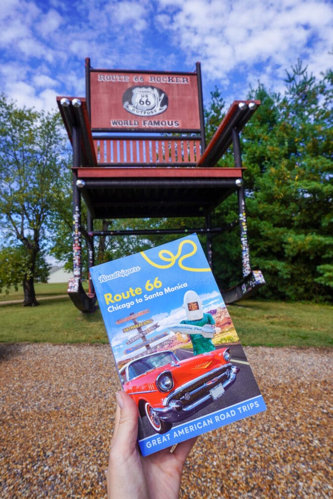A handing holding up a "Roadtrippers Route 66" guidebook in front of a very large rocking chair