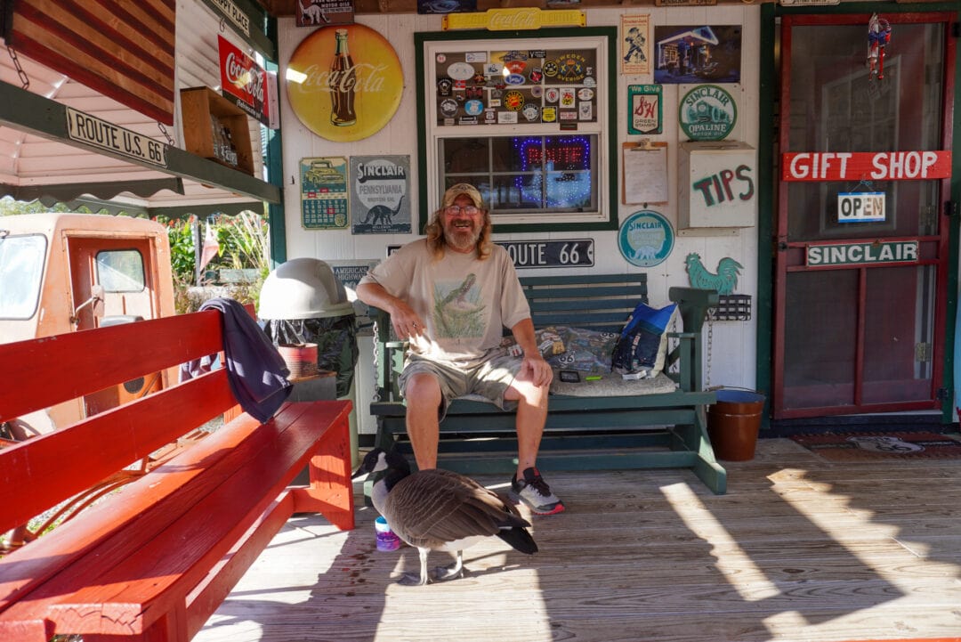 A smiling man seated on a bench on a porch next to a goose, surrounded by Route 66 memorabilia