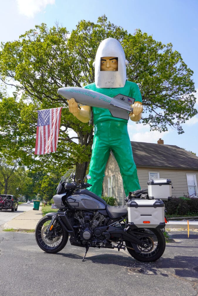 A motorcycle parked in front of a large fiberglass statue of a man holding a rocket ship
