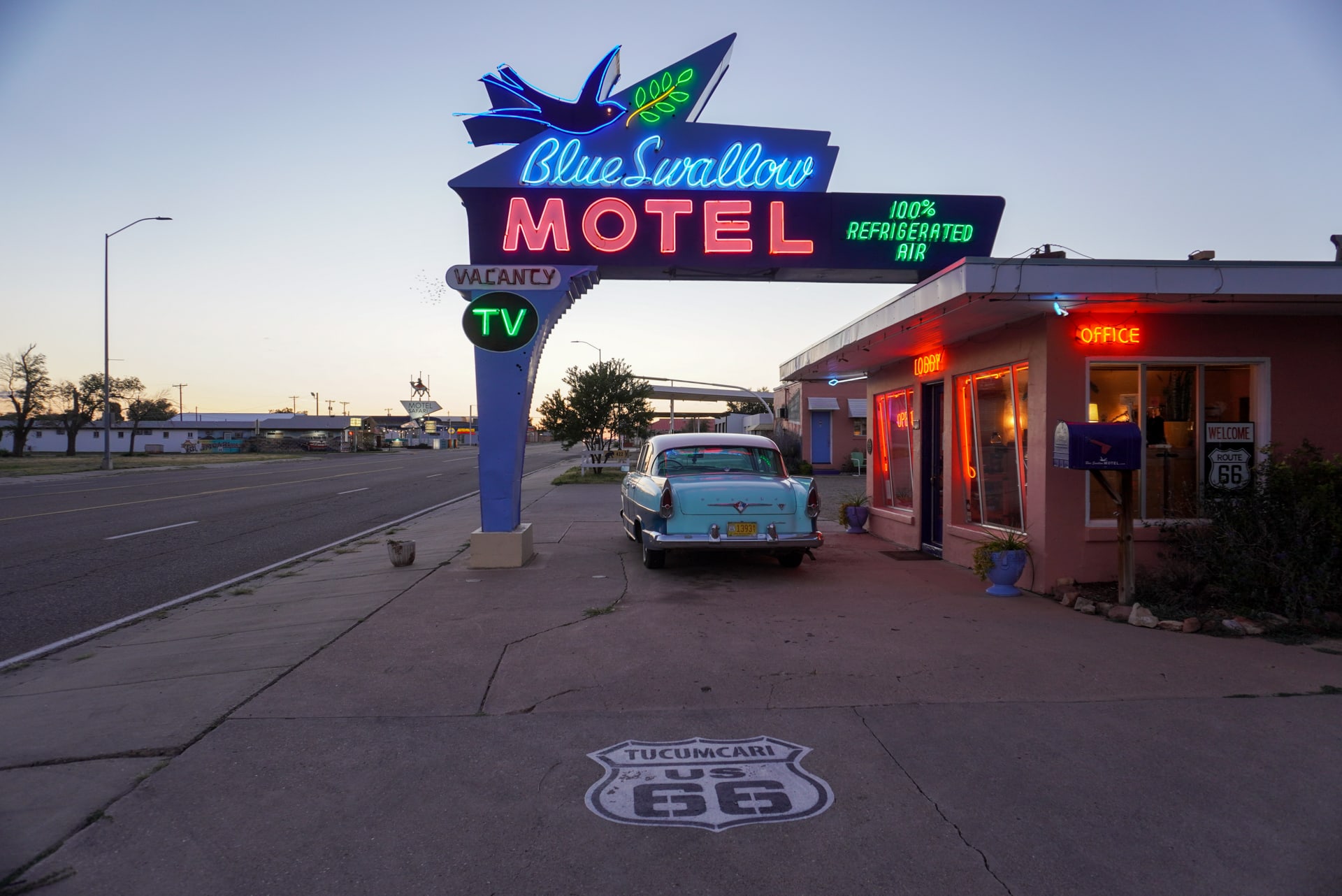 Exterior of a vintage motel at dusk, with a lit neon sign and a 1950s car parked at the entrance