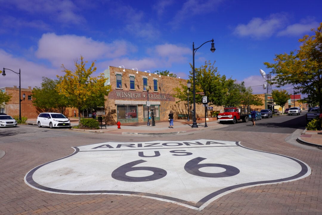 A large Arizona Route 66 shield painted on the ground in the middle of an intersection, with a park in the background