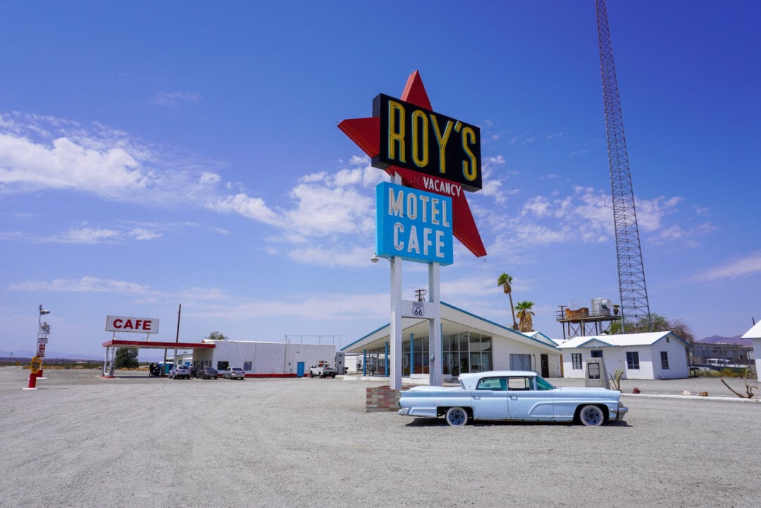 A light blue 1950s car is parked below a tall neon sign that reads "Roy's Motel Cafe, Vacancy"