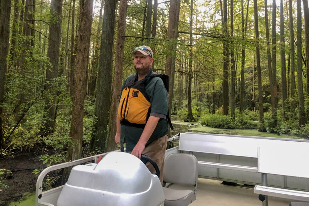 A man in a life jacket and baseball hat guides a white pontoon boat through a dense forest