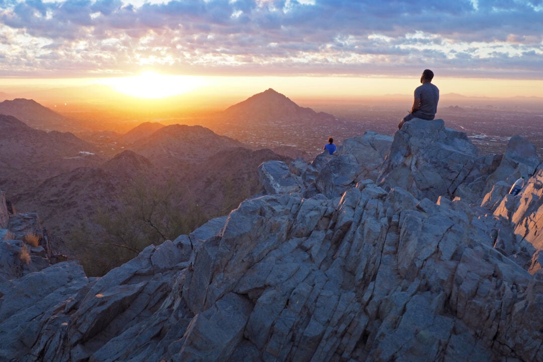 two people sit on tall rock formations and look out at a sunrise
