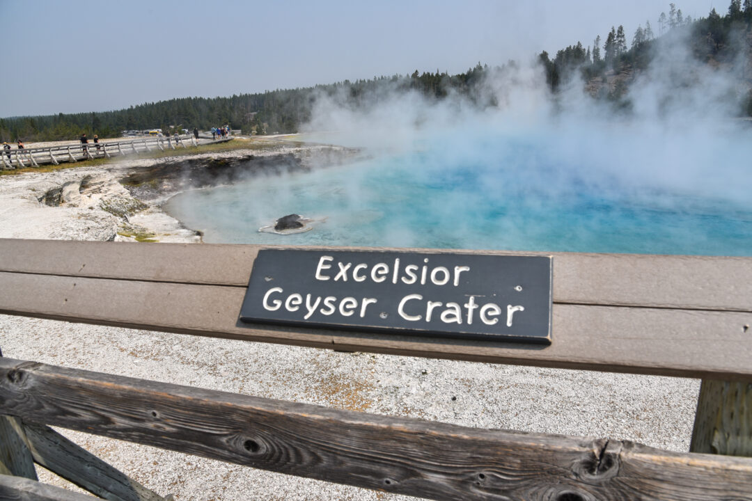 a wooden railing says "Excelsior Geyser Crater" in front of a steaming blue geyser