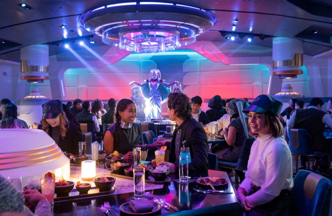 People gathered around a bar and dining tables in a futuristic restaurant with neon lights and a masked performer on stage