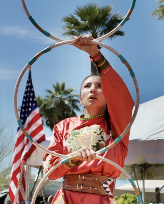 a woman in native american regalia holds three hoops in front of an american flag and blue sky