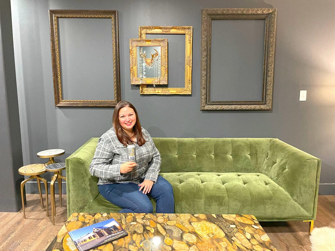 a woman sits on a green velvet couch holding a can of beer in front of a gray wall with gold frames