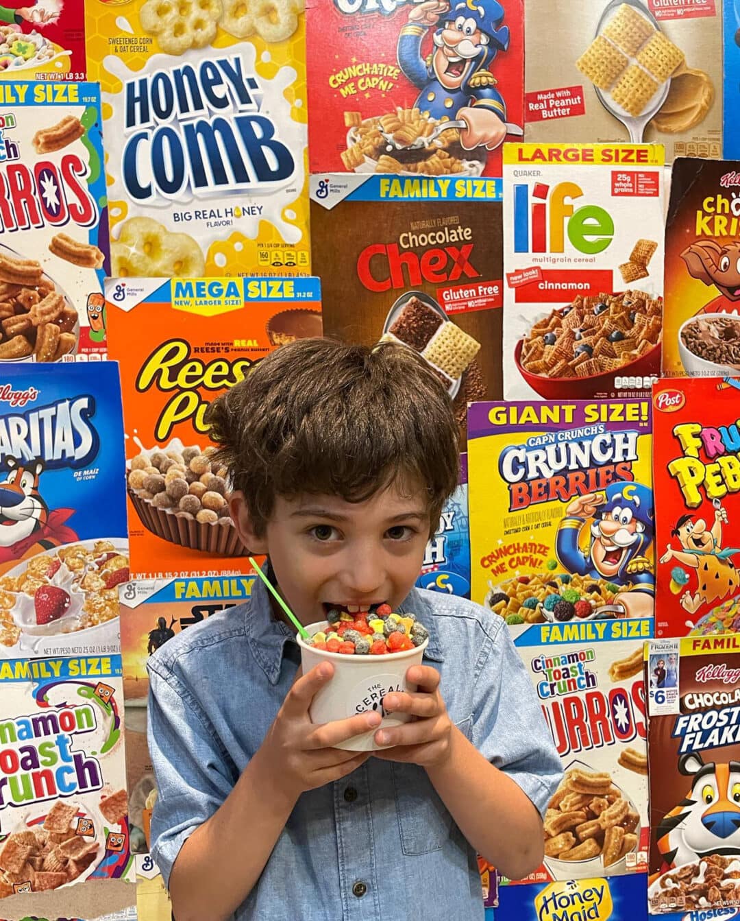 a boy stands eating a cup of cereal in front of a wall of cereal boxes