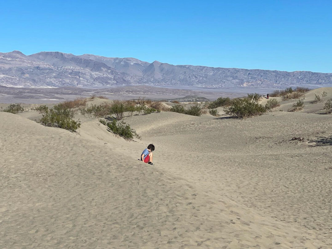 A small child sitting down in sand dunes with mountains in the background