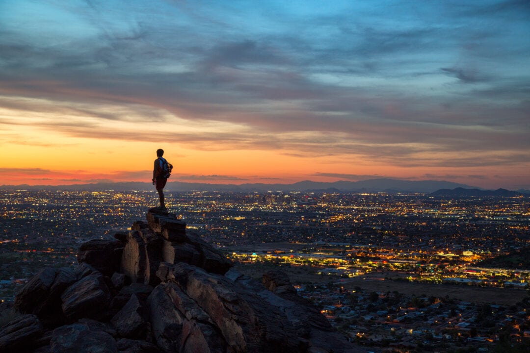 a person stands on a high rock formation overlooking a city at sunset