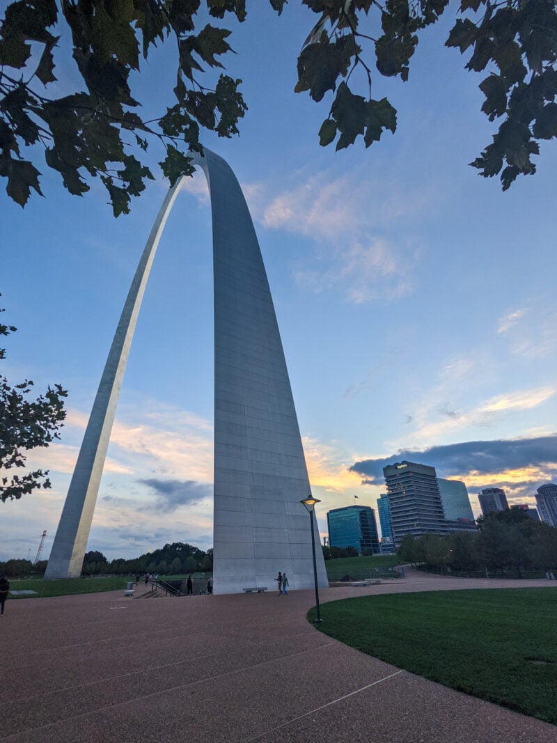 Very large arch structure with some greenery in the foreground and a city skyline and sunset in the background