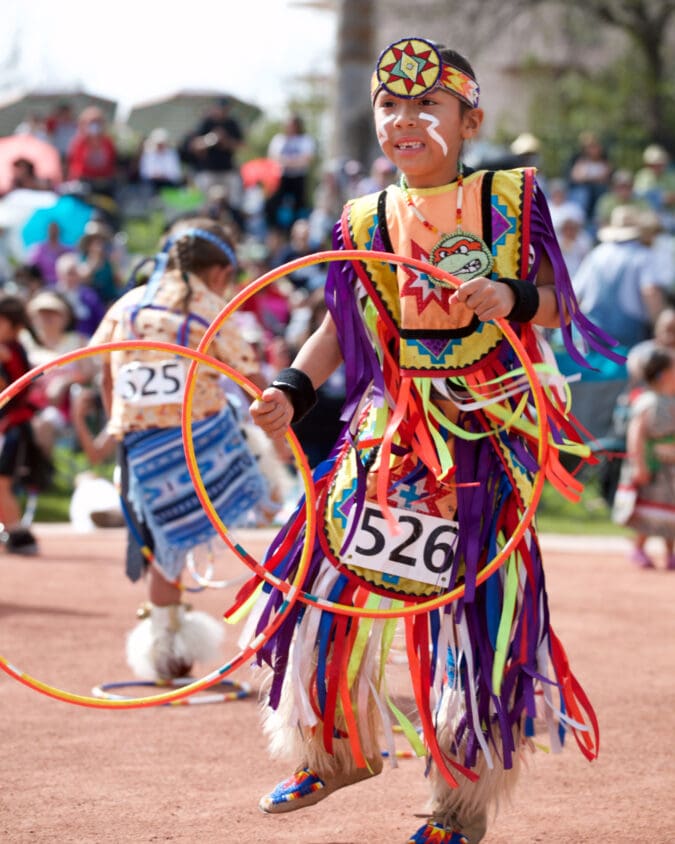 a boy in native regalia holds two orange hoops and dances in front of a crowd