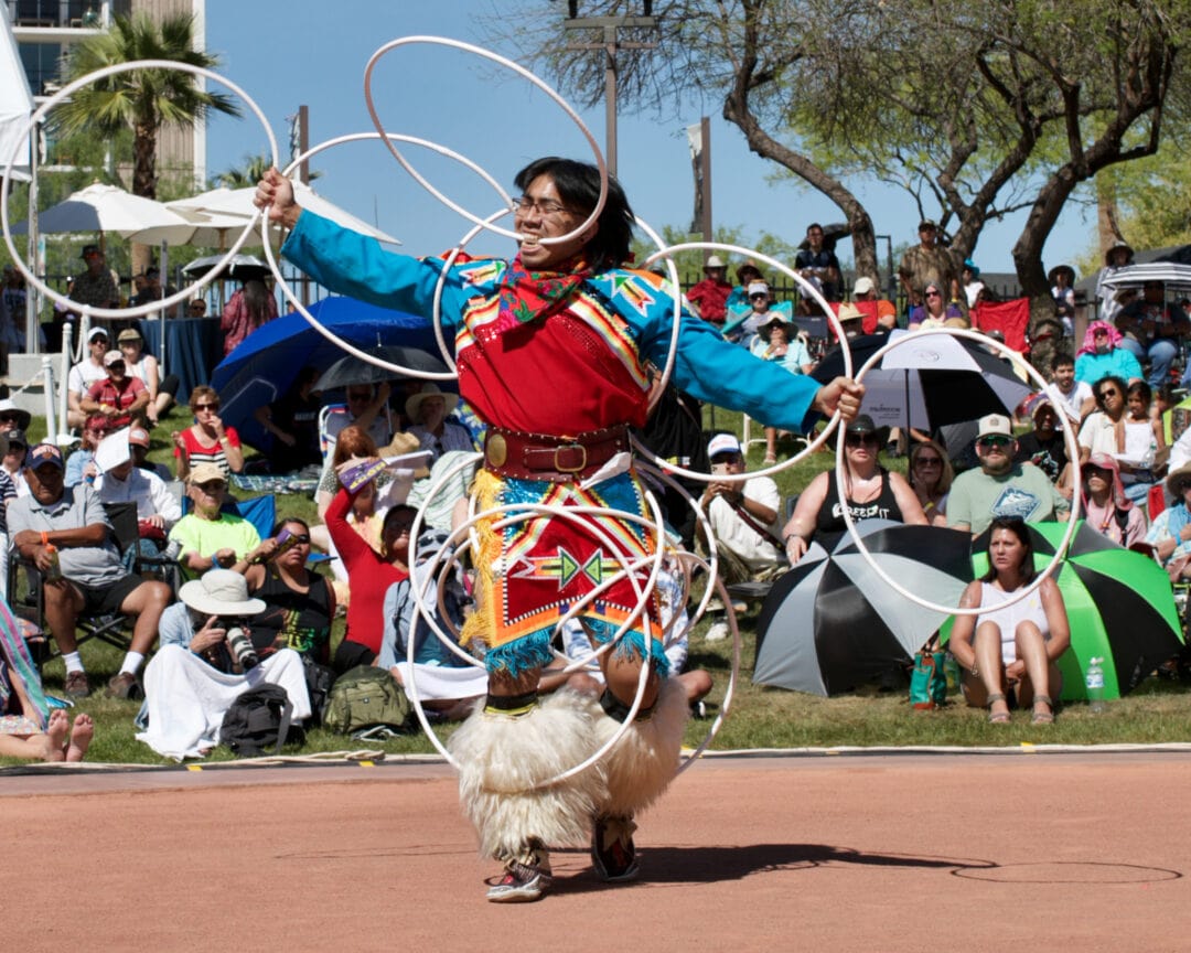 a man in native regalia has several hoops on his body including one in his mouth and dances in front of a crowd