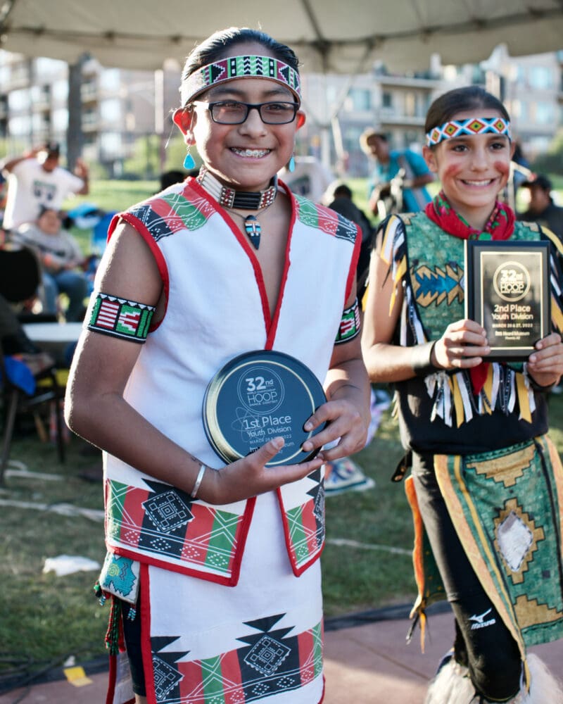 two boys in native american regalia hold awards and smile