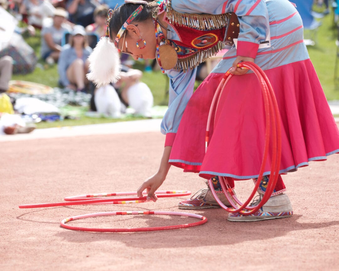 a woman in native regalia bends down to pick up a pink hoop while her other hand holds hoops