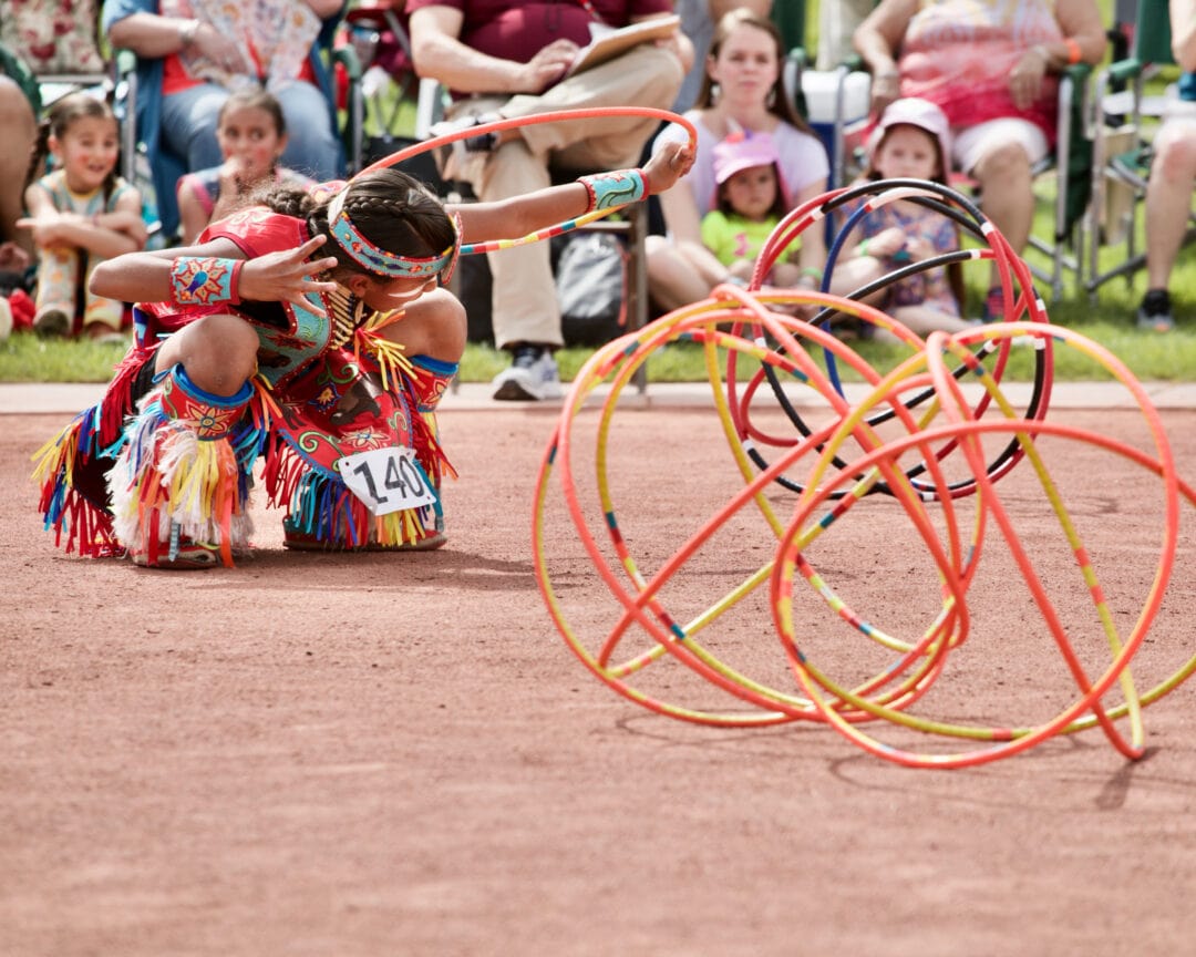 a boy in native american regalia holds an orange hoop next to a pile of orange hoops stacked