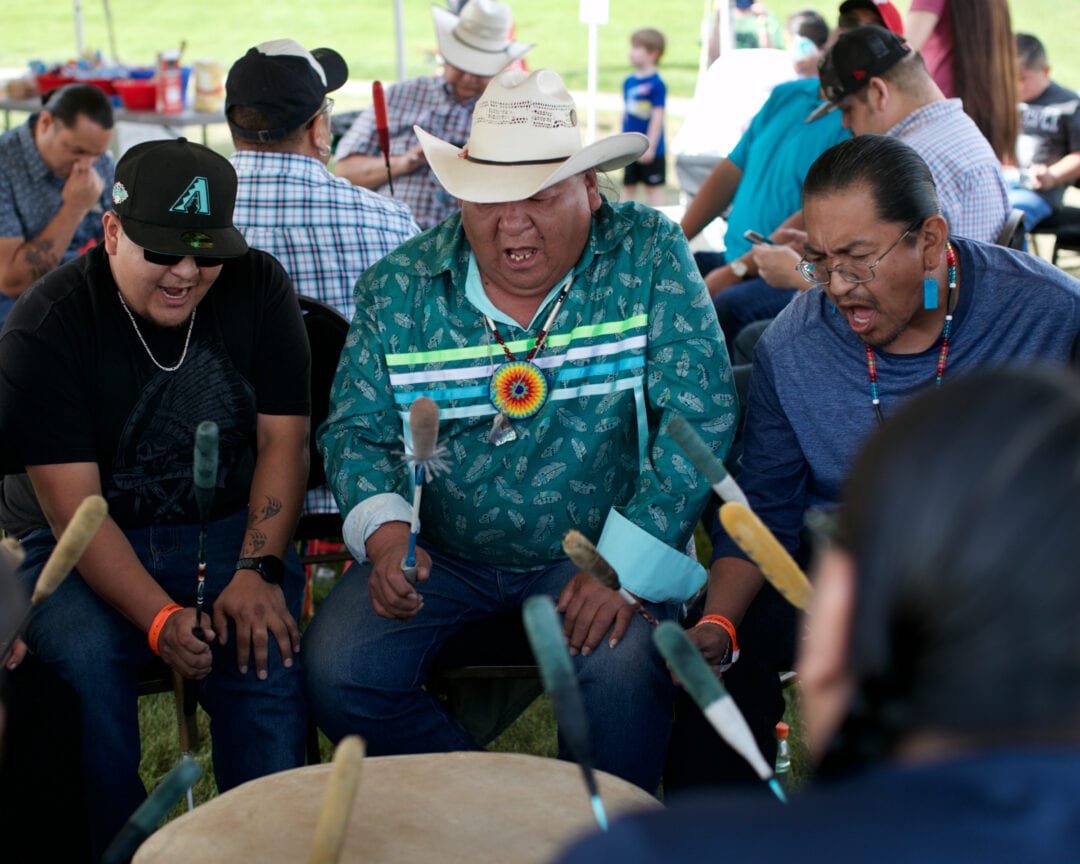 three men beat a drum and sing in a circle