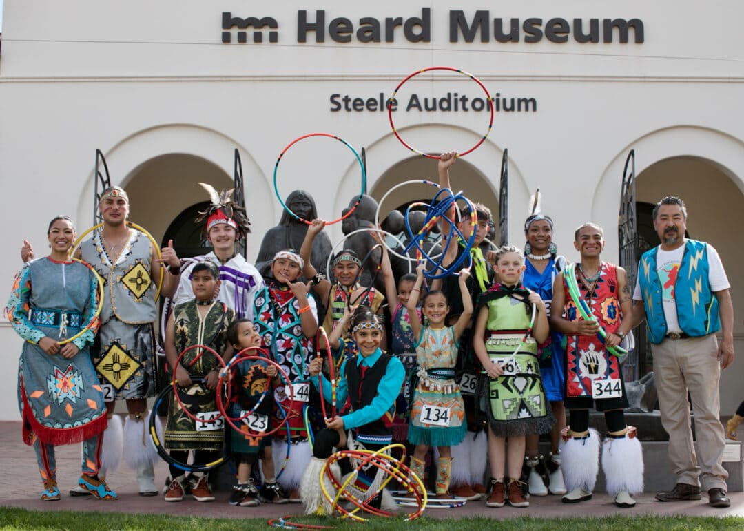 a group of children and adults pose in front of the heard museum in native regalia holding hoops