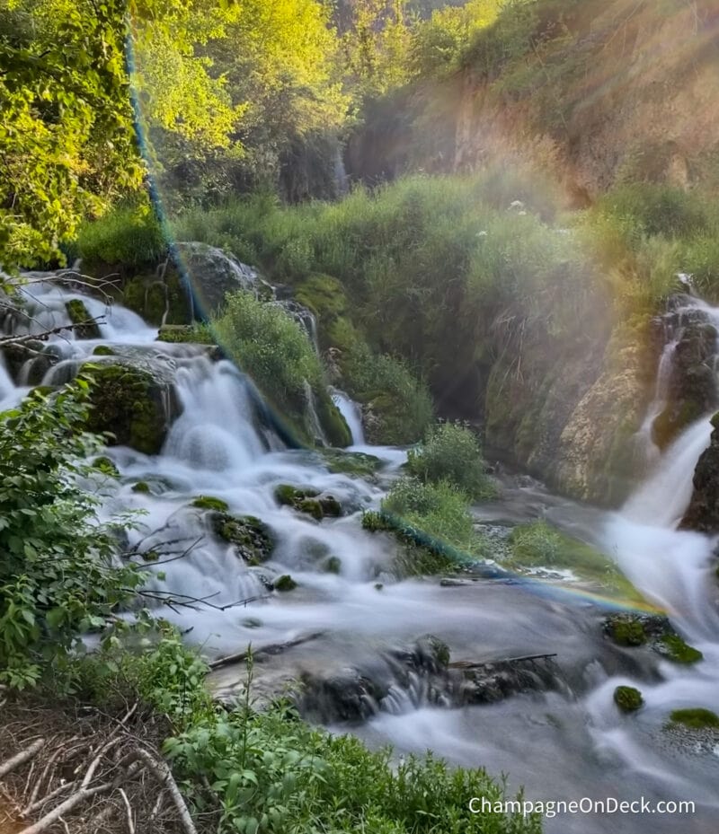 a long exposure photo of a waterfall in a forest with a sun flare and a rainbow